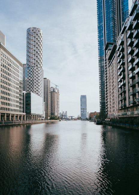 A wide view of a city canal in Canary Wharf, showing modern high-rise office buildings on both sides with reflective glass facades, some with visible balconies. The calm water in the canal displays ge