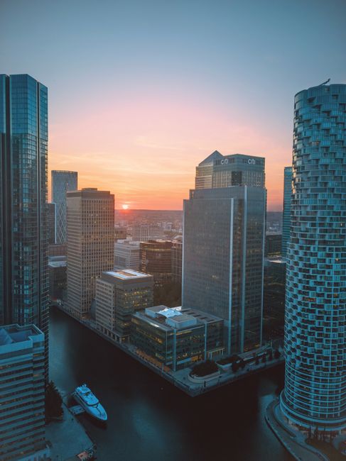 A wide view of a city canal in Canary Wharf, showing modern high-rise office buildings on both sides with reflective glass facades, some with visible balconies. The calm water in the canal displays ge