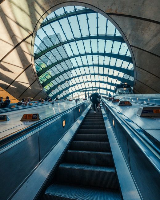 View of an indoor escalator ascending towards a large, arched glass roof structure in a modern building, with sunlight streaming through the glass panels. The escalator has stainless steel side panels