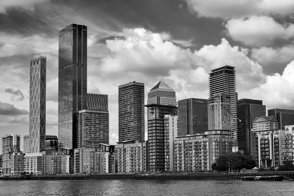 A cityscape of Marsh Wall flats in Canary Wharf showing several modern high-rise office buildings with glass facades reflecting the sky and surrounding structures. The tallest building has a distincti